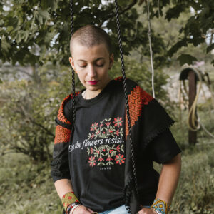 A person with a shaved head sits on a swing under a tree, wearing a black shirt with floral designs and text, light blue jeans, and colorful bracelets. A patterned shawl is draped over their shoulders. The background is lush and green.