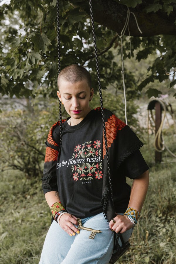 A person with a shaved head sits on a swing under a tree, wearing a black shirt with floral designs and text, light blue jeans, and colorful bracelets. A patterned shawl is draped over their shoulders. The background is lush and green.