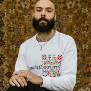 A bearded person sits cross-legged against an ornate, floral-patterned tapestry. They wear a white long-sleeve shirt with embroidered red flowers and the phrase "Even the flowers resist.