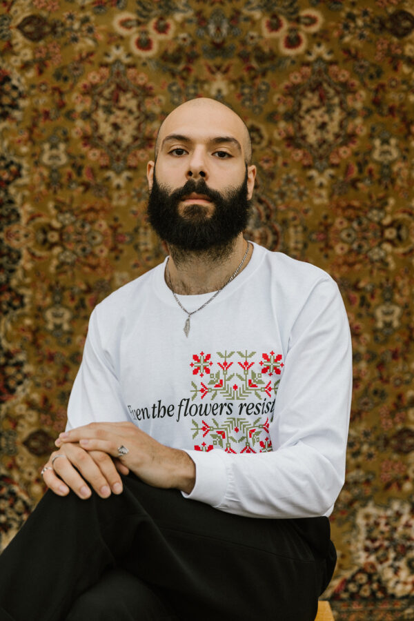 A bearded person sits cross-legged against an ornate, floral-patterned tapestry. They wear a white long-sleeve shirt with embroidered red flowers and the phrase "Even the flowers resist.