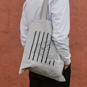 A person with a beard wearing a light gray long-sleeve shirt and black pants stands against a reddish wall. They carry a gray tote bag with black vertical lines and a geometric pattern, featuring the text "LIBERATION FOR ALL.