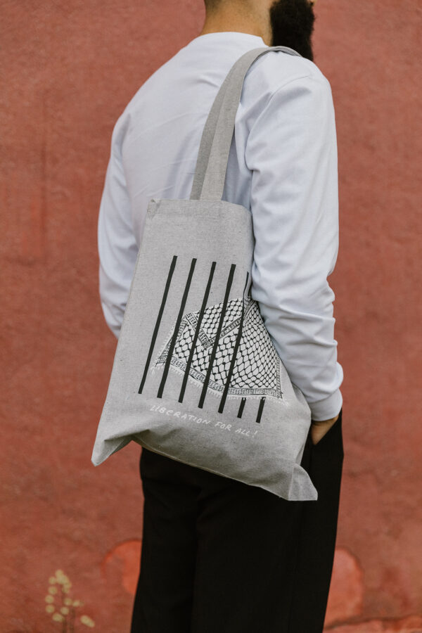 A person with a beard wearing a light gray long-sleeve shirt and black pants stands against a reddish wall. They carry a gray tote bag with black vertical lines and a geometric pattern, featuring the text "LIBERATION FOR ALL.