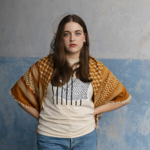 A woman with long brown hair stands with her hands on her hips. She wears a graphic t-shirt, blue jeans, and a patterned shawl over her shoulders. The background is a light blue, textured wall.