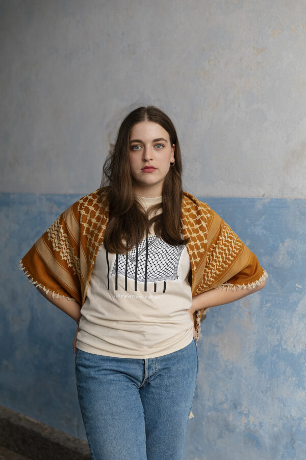 A woman with long brown hair stands with her hands on her hips. She wears a graphic t-shirt, blue jeans, and a patterned shawl over her shoulders. The background is a light blue, textured wall.