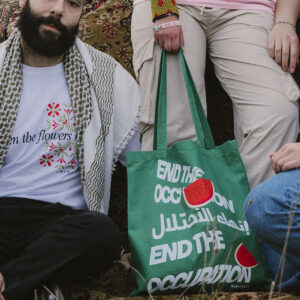 Two people are sitting side by side on a patterned rug. The person on the left wears a white shirt with floral design and a keffiyeh. The person on the right holds a green tote bag with "End the Occupation" printed in white in English and Arabic.