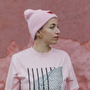 Person wearing a pink T-shirt with black and white graphic print and a pink beanie, standing against a textured pink wall. They have short hair and visible earrings.