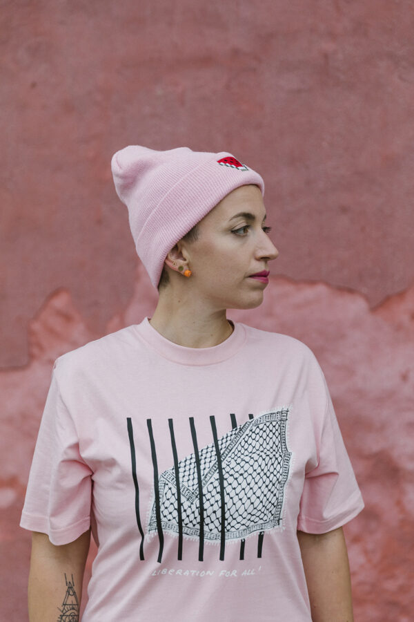 Person wearing a pink T-shirt with black and white graphic print and a pink beanie, standing against a textured pink wall. They have short hair and visible earrings.