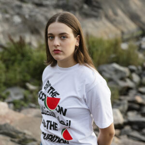 A person with long hair stands outdoors in front of a rocky landscape. They are wearing a white T-shirt with graphic text and images of watermelons, and blue jeans. The backdrop includes grass and rocks.