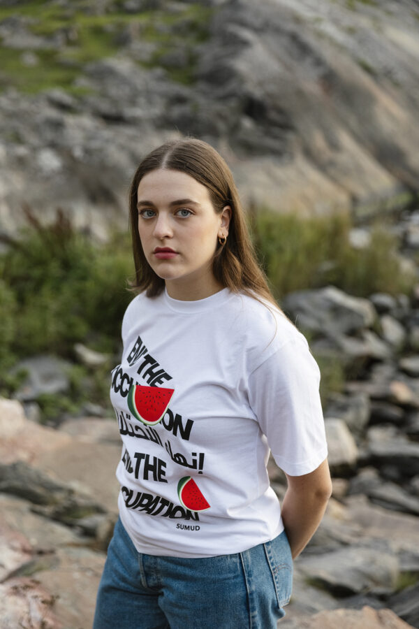 A person with long hair stands outdoors in front of a rocky landscape. They are wearing a white T-shirt with graphic text and images of watermelons, and blue jeans. The backdrop includes grass and rocks.