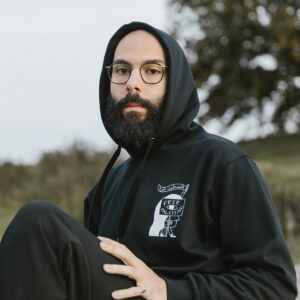 A person with a beard and glasses is sitting outdoors wearing a black hoodie with a design on the front. The background features trees and a cloudy sky. The person has one knee up and looks towards the camera.