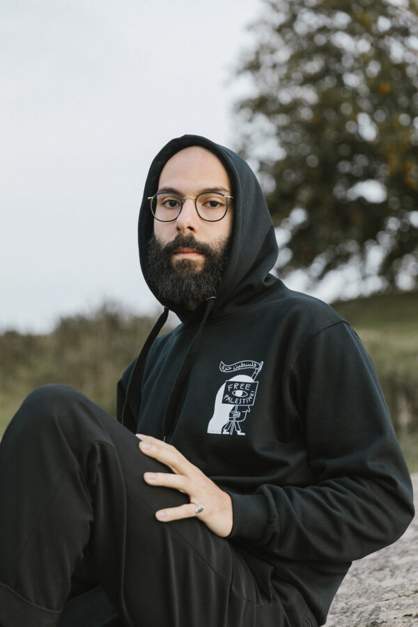 A person with a beard and glasses is sitting outdoors wearing a black hoodie with a design on the front. The background features trees and a cloudy sky. The person has one knee up and looks towards the camera.