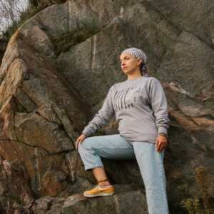 A person wearing a gray sweatshirt, checked bandana, light blue jeans, and yellow shoes stands on a rocky surface. They gaze into the distance with one foot raised on a rock, surrounded by stone and sparse vegetation.