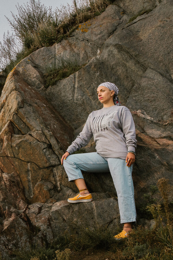 A person wearing a gray sweatshirt, checked bandana, light blue jeans, and yellow shoes stands on a rocky surface. They gaze into the distance with one foot raised on a rock, surrounded by stone and sparse vegetation.