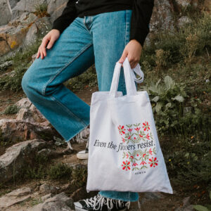 A person stands on rocky steps, wearing jeans and black sneakers, holding a white tote bag with floral designs and the text "Even the flowers resist." The background features rocks and greenery.