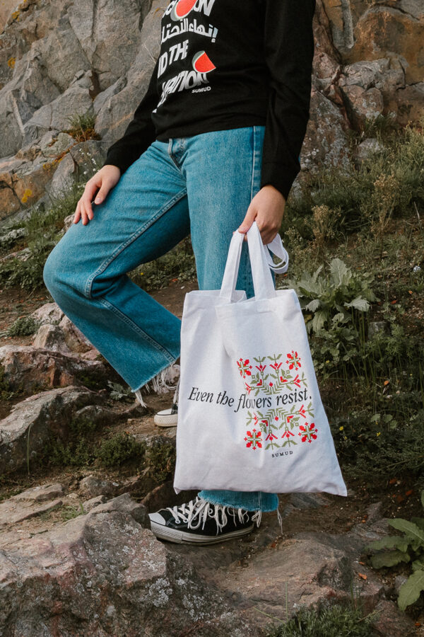 A person stands on rocky steps, wearing jeans and black sneakers, holding a white tote bag with floral designs and the text "Even the flowers resist." The background features rocks and greenery.