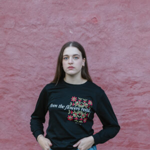 A person with long hair stands against a textured pink wall. They wear a black long-sleeve shirt with floral designs and the text "even the flowers resist." They have their hands in their jean pockets and look towards the camera.