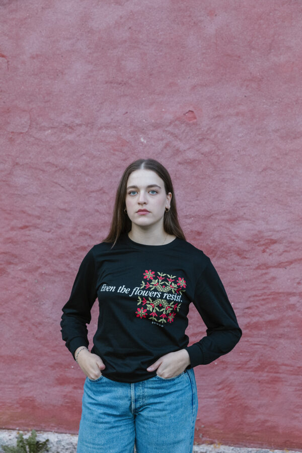 A person with long hair stands against a textured pink wall. They wear a black long-sleeve shirt with floral designs and the text "even the flowers resist." They have their hands in their jean pockets and look towards the camera.