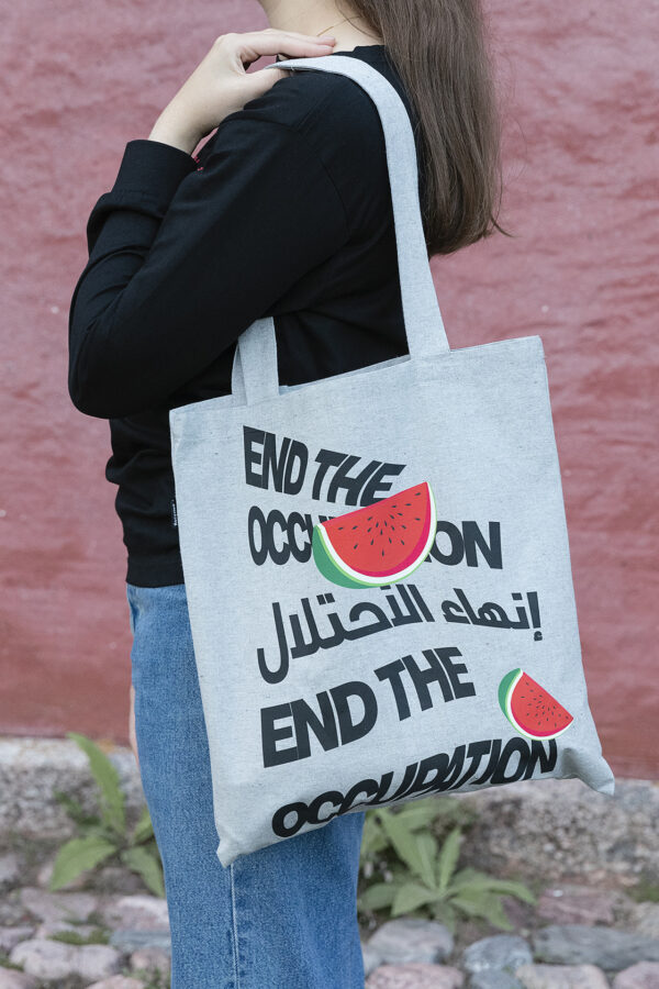 A person holds a tote bag with the text "End the Occupation" in English and Arabic, along with images of watermelon slices. The person is wearing a black top and jeans, standing against a red wall.