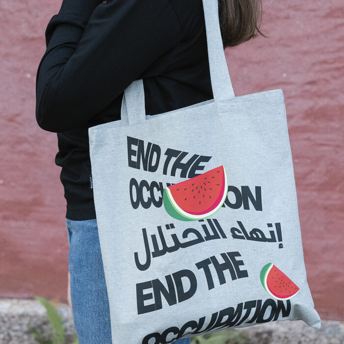 A person holds a tote bag with the text "End the Occupation" in English and Arabic, along with images of watermelon slices. The person is wearing a black top and jeans, standing against a red wall.