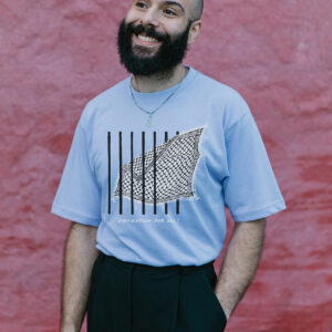 A bearded man smiles while standing against a textured red wall. He wears a light blue t-shirt with a graphic design and black pants. He has a shaved head and a necklace.