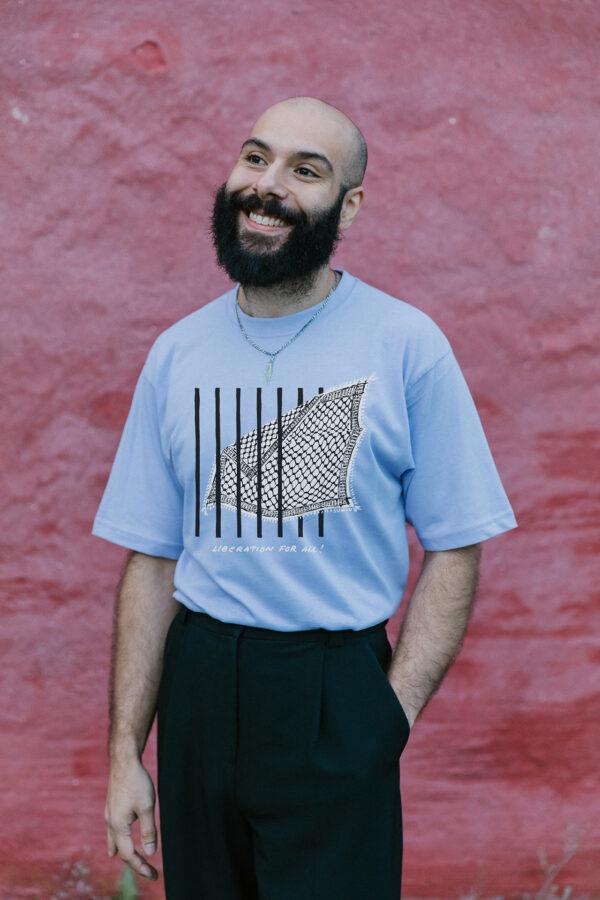 A bearded man smiles while standing against a textured red wall. He wears a light blue t-shirt with a graphic design and black pants. He has a shaved head and a necklace.