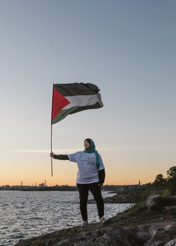 A person stands on a rocky shore at dusk, holding a Palestinian flag. They wear a light blue hijab and a white shirt. The sky is clear and the calm sea stretches in the background.