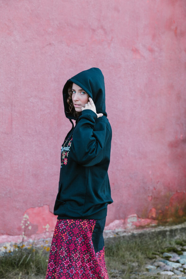 A person in a black hoodie and a red patterned skirt stands outdoors against a pink wall, looking sideways at the camera. There's grass and stones on the ground.