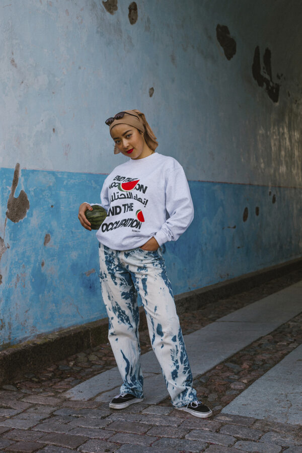 A person stands on a cobblestone path against a blue and white wall. They wear sunglasses on their head, a white sweatshirt with text, bleached jeans, and black shoes, holding an avocado in one hand.