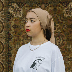 A woman wearing a beige headscarf, red lipstick, and a white long-sleeve shirt with a design stands in front of a detailed, patterned rug.