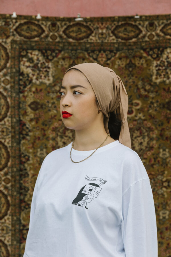 A woman wearing a beige headscarf, red lipstick, and a white long-sleeve shirt with a design stands in front of a detailed, patterned rug.