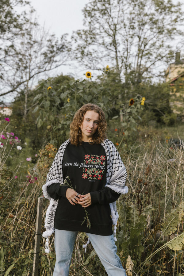 A person with curly hair stands outdoors in front of greenery and sunflowers, wearing a black sweater with floral designs and text, and a patterned shawl draped over their shoulders, holding a small flower.