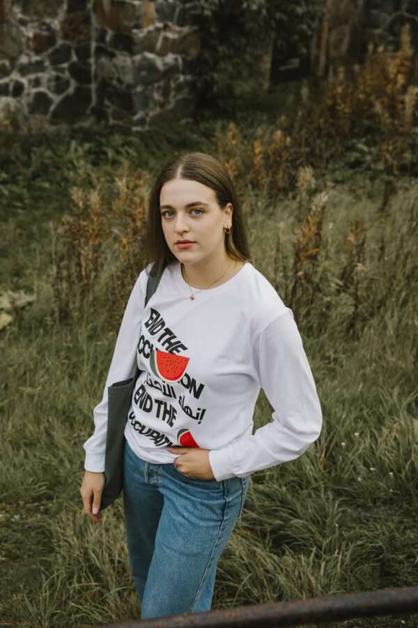 Young woman with long hair stands outdoors on grass, wearing a white long-sleeve shirt with black text and a red graphic, and blue jeans. There's a stone wall and tall plants in the background.