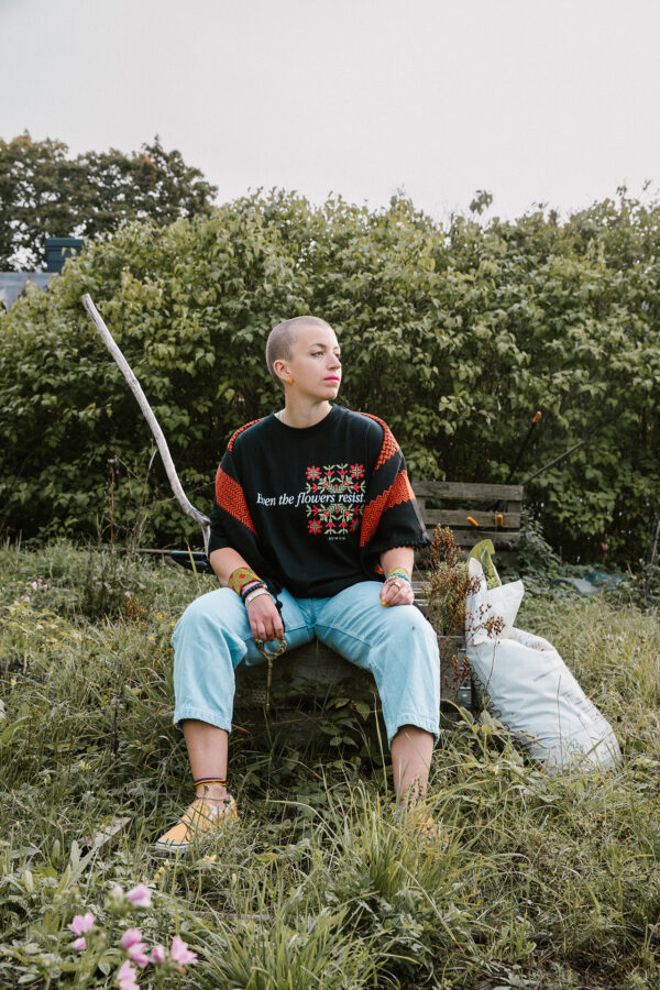 Person with a shaved head sits on a wooden bench in a garden, wearing a black sweater with red patterns and light blue jeans. Green foliage is in the background. A white bag is on the ground nearby.