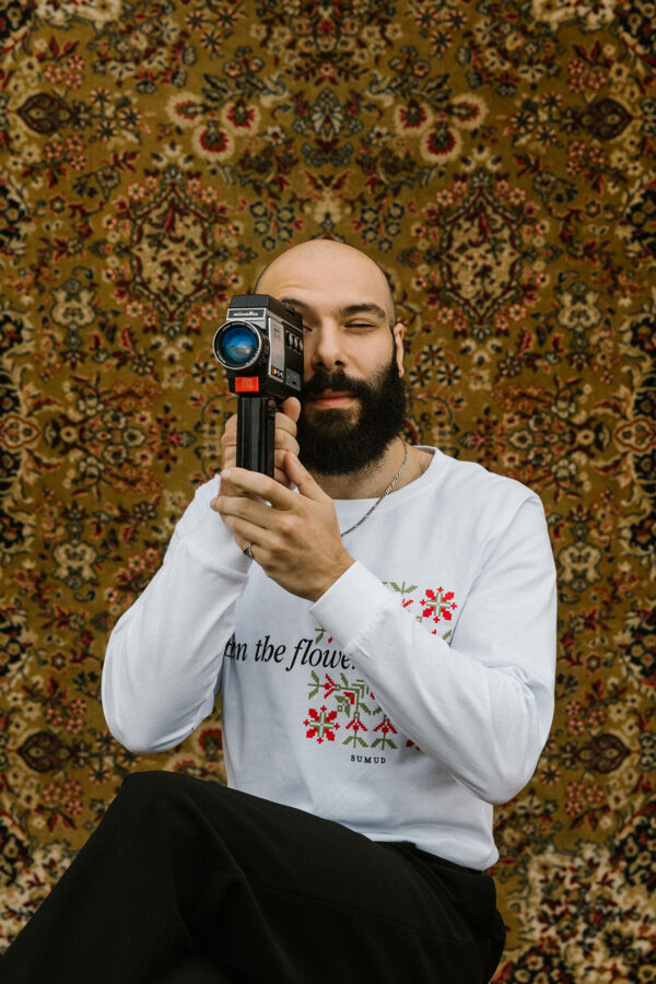 A bearded man holds a vintage camera while sitting against an ornate floral-patterned tapestry. He wears a white sweatshirt with red flower designs, and black pants. The background creates a warm and artistic atmosphere.