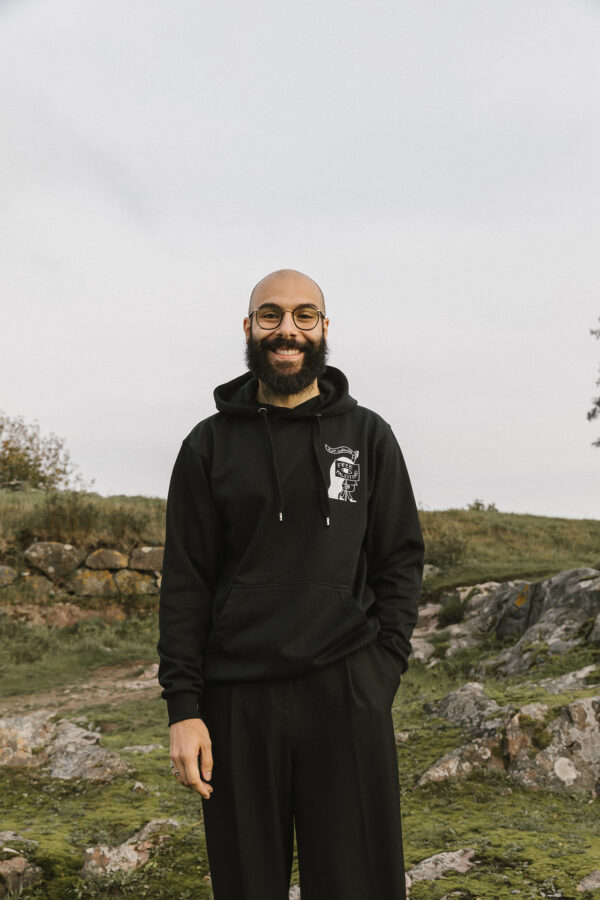 A smiling person with glasses and a beard stands outdoors on grassy, rocky terrain. They are wearing a black hoodie and pants. The background features overcast skies and scattered vegetation.