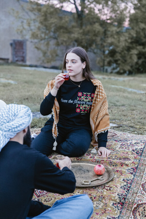 A woman sitting on a patterned rug outdoors, holding a piece of pomegranate to her lips. She's wrapped in a shawl and wears a floral shirt. A man sits nearby with a turban, and there's a wooden tray with a pomegranate on the rug.