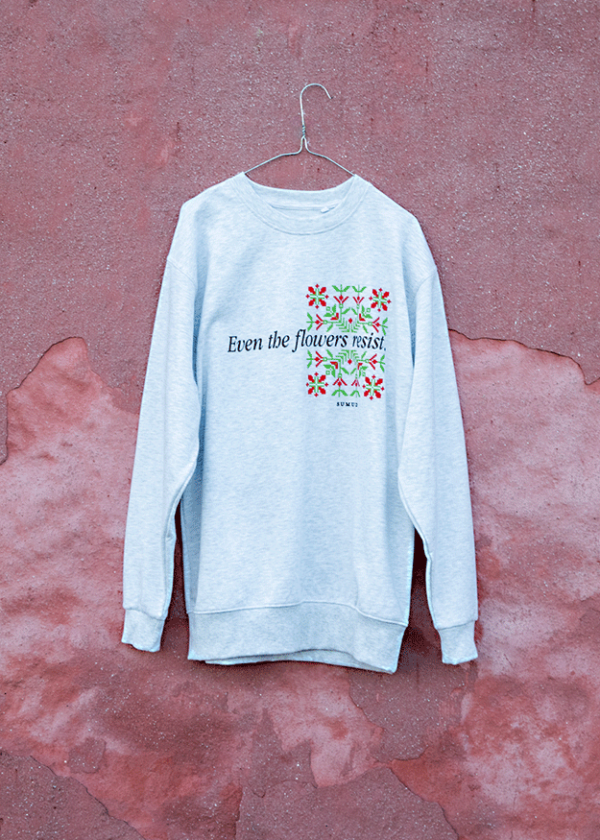 A light gray sweatshirt hanging on a hook against a textured pink background. The sweatshirt has floral graphics and the text "Even the flowers resist" printed on the front.