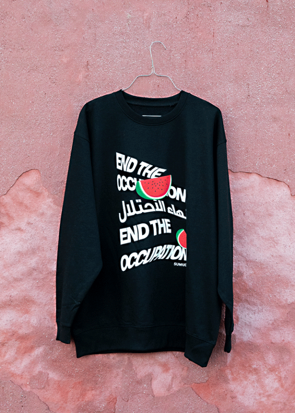 Black sweatshirt hanging on a wire against a textured pink wall. The shirt features the phrase "End the Occupation" in English and Arabic, with stylized watermelon graphics.