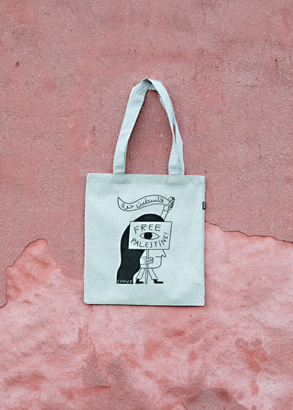 A tote bag hangs against a textured pink wall. The bag features a drawing of a person holding a sign that reads "Free Palestine," with a banner above saying "Be Vibrant.