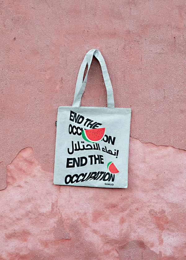 A gray tote bag hanging against a textured pink wall. The bag features the text "End the Occupation" in English and Arabic, with illustrated watermelon graphics.