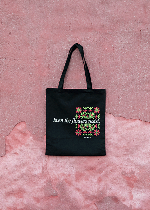 A black tote bag with floral designs and the text "Even the flowers resist" printed on it. The bag is laid flat on a textured, pinkish surface.