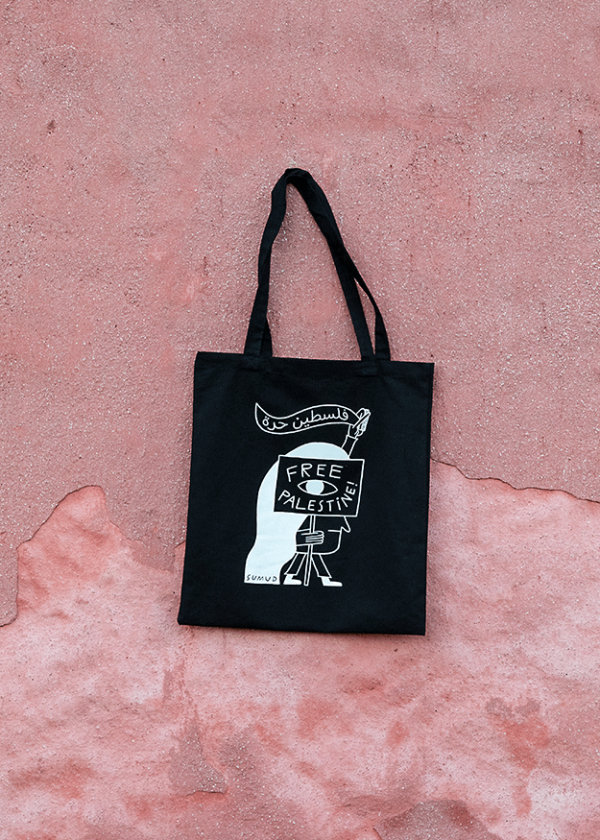 A black tote bag with a stylized illustration and the text "Free Palestine" hangs against a textured pink wall.