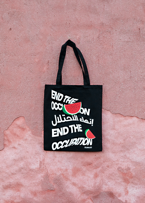 A black tote bag hangs against a textured pink wall. It features the text "END THE OCCUPATION" with watermelon graphics, and Arabic script between repeated phrases.