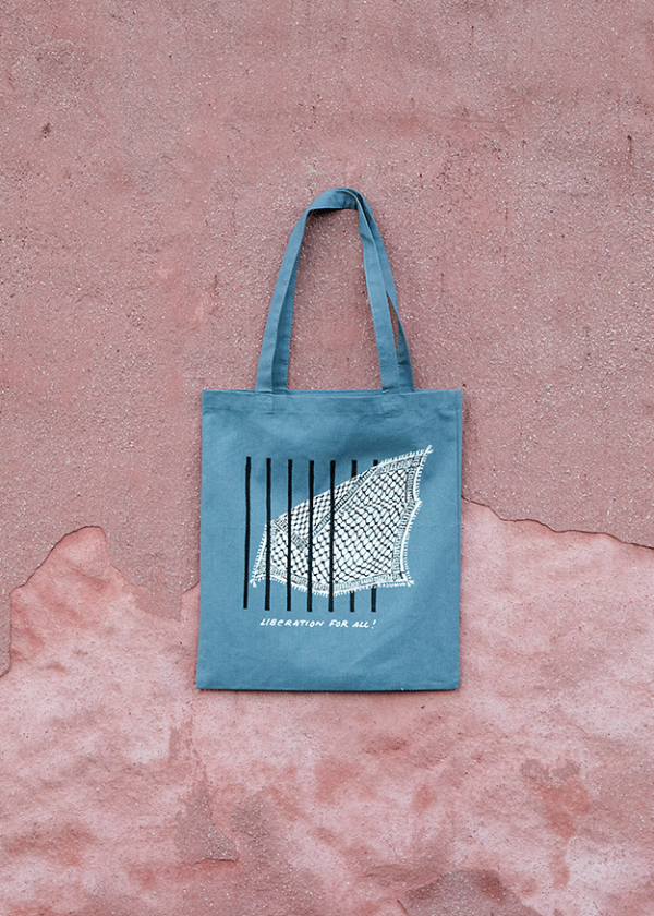 A blue tote bag with an illustration of a bird netted behind bars. The text "Liberation For All" is below the design. The bag is hanging against a textured, pinkish wall.