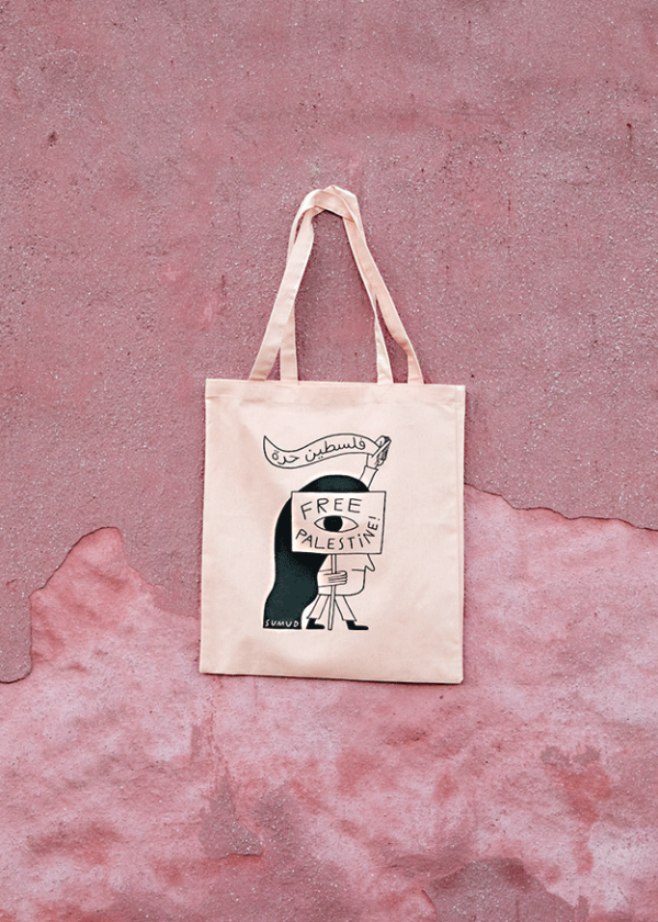 A beige tote bag with an illustration of a person holding a sign that reads "Free Palestine" in black text. The background is a textured pink surface.