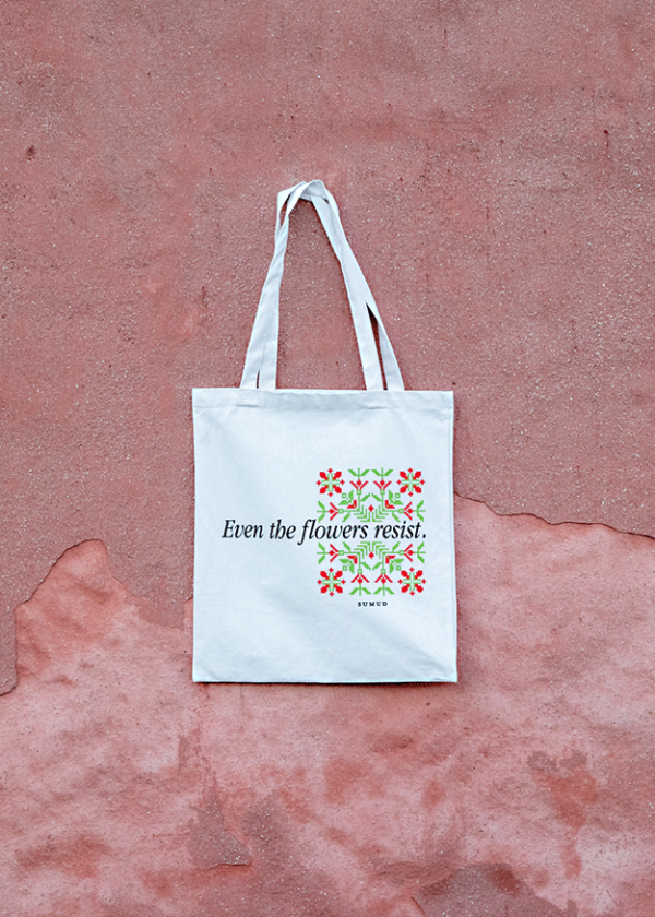 A white tote bag hangs against a textured pink wall. The bag features a floral design with the text "Even the flowers resist.