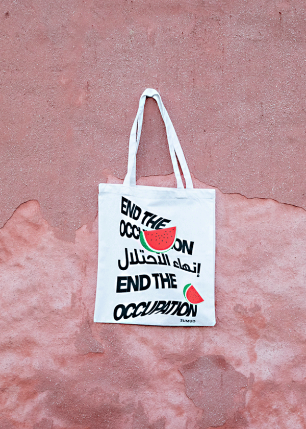 A white tote bag with the phrase "End the Occupation" written in wavy black letters. The design includes an image of a watermelon slice. The background is a textured, pale pink surface.