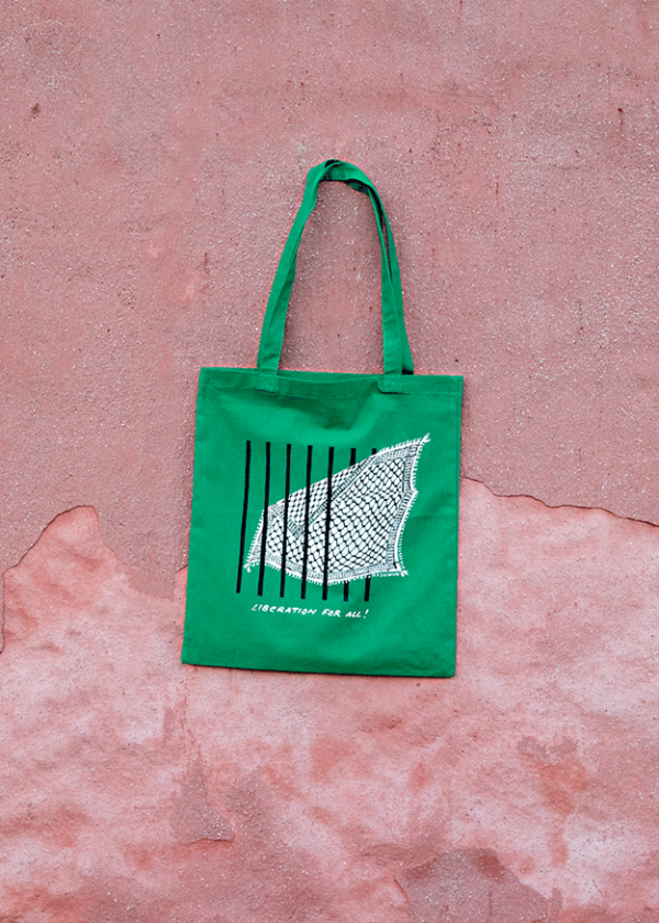 A green tote bag hangs against a textured pink wall. The bag features a graphic of a bird wing with black vertical lines resembling bars and the text "LIBERATION FOR ALL!" printed below the design.