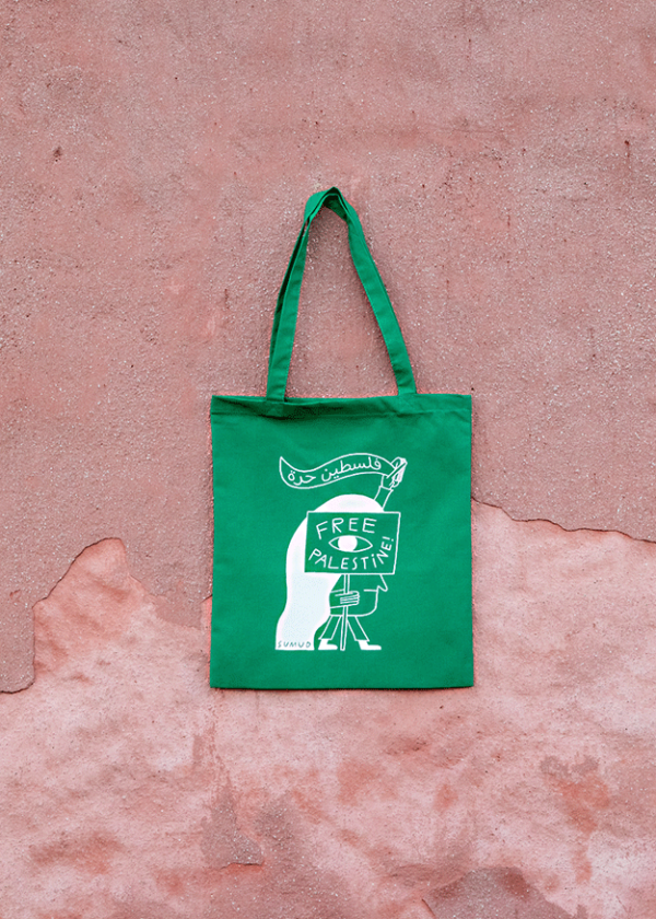 A green tote bag with a graphic of a figure holding a sign that reads "Free Palestine" in white. The bag is hanging against a textured, pinkish wall.