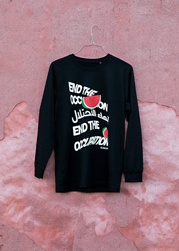 A black long-sleeve shirt hangs against a textured, pink wall. The shirt features white text reading "END THE OCCUPATION" in English and Arabic, with small images of watermelon slices.